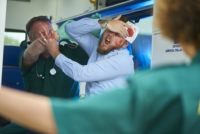 two ambulance paramedics stabilise an assault victim. They are wearing green ambulance uniform typical of uk paramedics. One is sitting in the back of the car stabilising the victim's head whilst the other paramedic is closing the door of the ambulance . The victim has been out drinking and has either fallen or been in a fight , and is acting aggressively towards the paramedic.